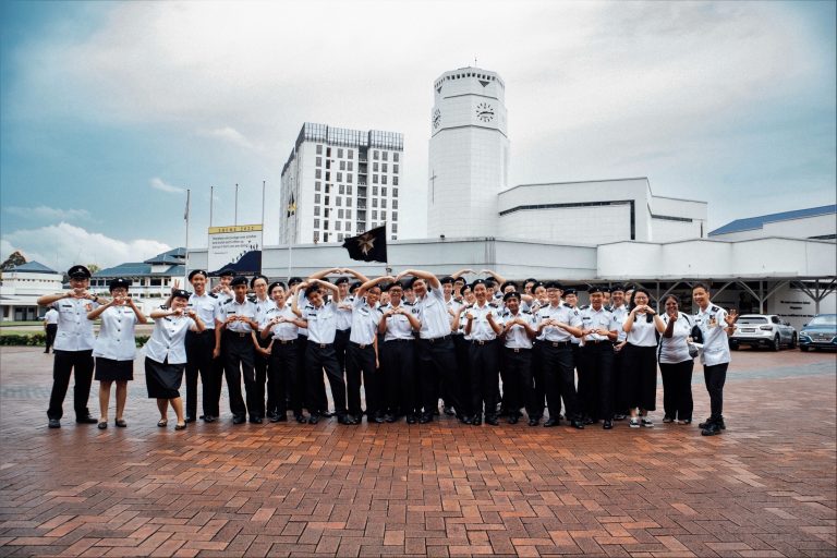 A large group of ACS(I) St John's Brigade members poses in white uniforms in front of the school clock tower.